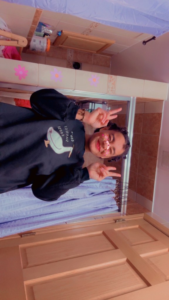 angel smiling with their eyes closed and holding two peace signs up with their hands; no glasses on her face and a black sweatshirt that reads God's silliest goose!